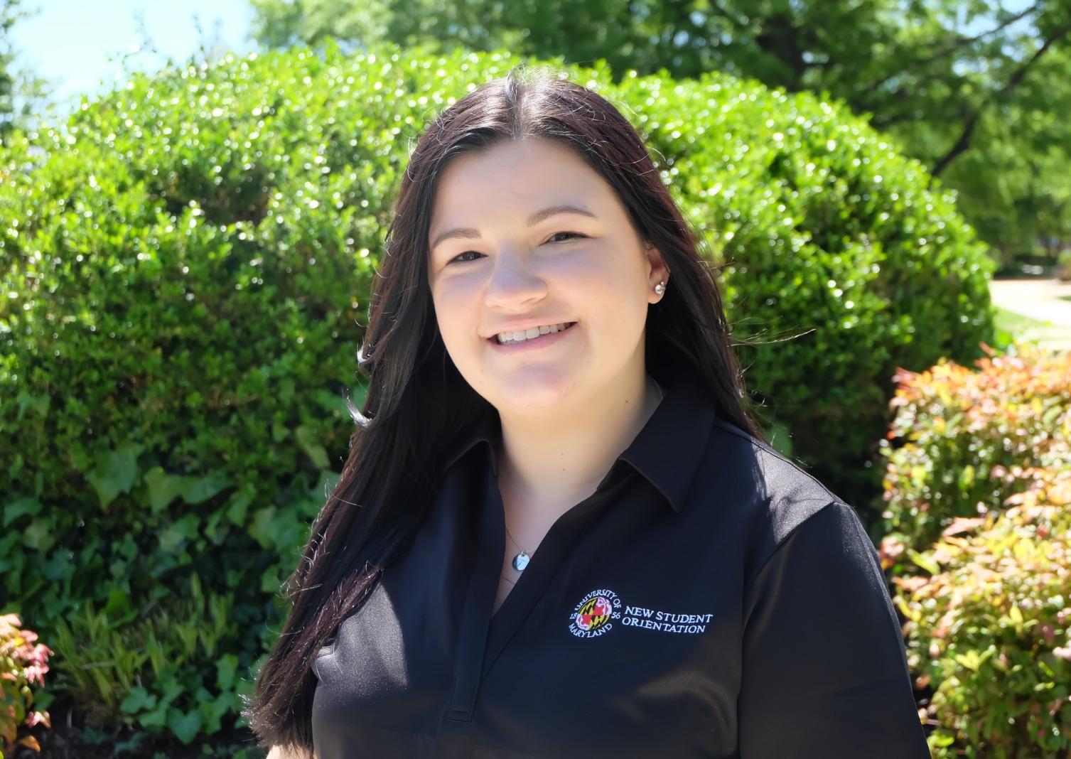 Tori is pictured in a black New Student Orientation and Transition polo in front of some trees on UMD's campus.