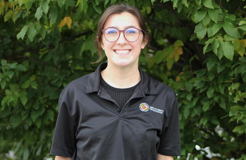 Meghan is pictured in a black New Student Orientation and Transition polo in front of some trees on UMD's campus.
